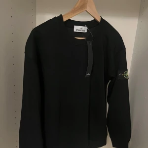Svart sweatshirt från Stone Island - Svart sweatshirt från Stone Island med klassisk rund hals och ikonisk logotyp-patch på vänster ärm. Tröjan är i bomull och har en clean, stilren look med ribbade muddar vid ärmslut och nederkant. Perfekt för dig som gillar streetwear och vill ha något stilrent.