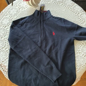 Mörkblå half zip tröja från Polo Ralph Lauren - Mörkblå långärmad tröja från Polo Ralph Lauren med röd broderad logga på bröstet. Tröjan har half zip-dragkedja och hög krage, tillverkad i mjuk bomull med diskret struktur. Perfekt för lager-på-lager och stilren look.