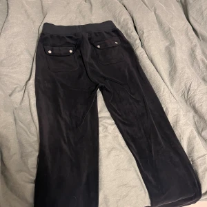 Svarta mjukisbyxor Juicy Couture XS - Svarta mjukisbyxor från Juicy Couture i storlek XS. Byxorna har bred resår i midjan med snörning, två fickor fram och två bakfickor med lock och silverfärgade knappar. Mjukt och skönt material, perfekt för chill dagar. Saknar en silvergrej på ena snöret, annars i mycket bra skick och bar använda ett fåtal gånger.