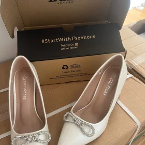 Vita pumps med rosett från CHC Shoes - Snygga vita pumps från CHC Shoes med spetsig tå och chunky klack. Skorna har en glittrig rosettdetalj framtill som ger extra bling. Perfekta för dig som vill sticka ut med stilrena och eleganta skor. Tillverkade i syntetmaterial med en klassisk form. Jag köpte från London på 1000kr 