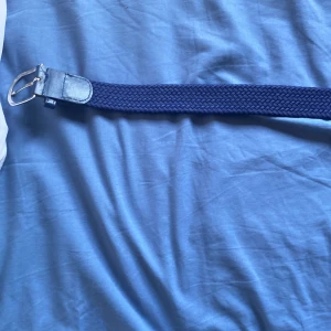 Mörkblått flätat bälte med skinn - Snyggt mörkblått bälte med flätad design och detaljer i skinn vid spännet och änden. Bältet har ett klassiskt silverfärgat spänne och passar perfekt till jeans eller chinos. Enkel och stilren look som funkar till många outfits.