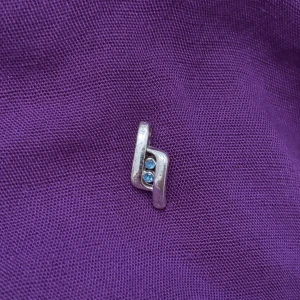 Silverfärgat hängsmycke med blå stenar - Unikt hängsmycke i silverfärg med modern, böjd form och två små blå stenar infällda i mitten. Perfekt att hänga på en kedja för en stilren och cool look. Passar dig som gillar minimalistiska detaljer och vill ha något som sticker ut.