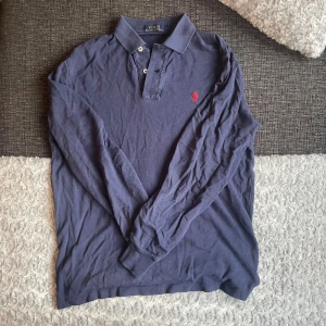 Blå långärmad pikétröja Polo Ralph Lauren - Klassisk marinblå långärmad pikétröja från Polo Ralph Lauren med röd broderad logga på bröstet. Tröjan har krage, knappar vid halsen och är gjord i mjuk bomull. Perfekt för en stilren och avslappnad look. Hör av er vid frågor och inga defekter på den⚡️☺️