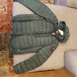 Grön dunjacka Parajumpers S - Snygg grön dunjacka från Parajumpers i storlek S. Jackan har huva, dragkedja framtill, två sidofickor med dragkedja och en patch på ärmen. Insidan är ljusbeige och jackan är quiltad för extra värme. Perfekt för kyliga dagar.