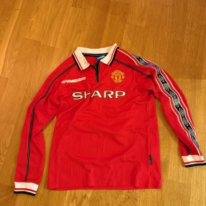 Retro Manchester United Beckham tröja Umbro S - Klassisk Manchester United långärmad fotbollströja från Umbro med Beckham 7 på ryggen. Röd med vita och svarta detaljer, klubbmärke på bröstet och SHARP som sponsor. Snygga mönster längs ärmarna och vit krage. 