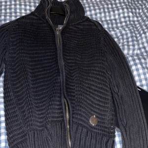 Svart stickad kofta från G-Star RAW - Säljer en svart, grovstickad kofta från G-Star RAW med dragkedja framtill och hög krage. Snygg broderad logga på ryggen och en metallbadge vid midjan. Perfekt för dig som gillar streetwear och vill ha något både stilrent och bekvämt.
