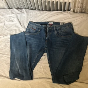 Blå Tommy Hilfiger Denim Slim Saber jeans - Säljer ett par blå Hilfiger Denim Slim Saber jeans med klassisk femficksdesign, snygga slitningar och diskreta röda detaljer vid fickorna. Jeansen har normal passform och raka ben, tillverkade i bomullsdenim med Hilfiger-läderpatch bak.