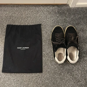 Svarta mockasneakers från Saint Laurent - Säljer ett par svarta sneakers från Saint Laurent i mocka med vita detaljer på hälen och beige sula. Skorna har klassisk snörning och låg profil, perfekt för en clean och stilren look. Medföljer original dustbag från Saint Laurent.