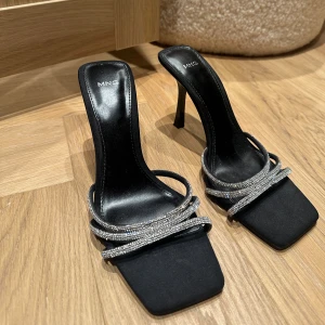 Svarta klackpumps med strass från MNG - Snygga svarta pumps med hög klack och fyrkantig tå från MNG. Skorna har flera tunna remmar dekorerade med glittrande strass som ger en lyxig vibe. Materialet är skinnimitation och designen är stilren och modern, perfekt för dig som gillar att sticka ut.