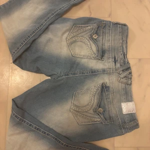 Ljusblå low waist bootcut jeans med slitningar och unika fickor - Ett par ljusblå bootcut jeans från Arya Jeans. De har slitna detaljer framtill, markerade sömmar och snygga broderade bakfickor med knapp. Det är denim och de har en tvättad look !!         Midjemått: 42cm , Yttreben: 99cm , Inreben: 70cm 