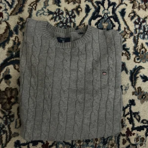 Grå kabelstickad tröja från Gant - Kabelstickad grå tröja från Gant med rund halsringning och diskret broderad logga på bröstet. Klassisk och stilren design i mjukt material, perfekt för lager-på-lager under kyligare dagar.