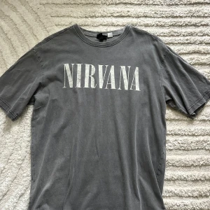 Grå Nirvana t-shirt med tryck - Snygg grå t-shirt med stort vitt Nirvana-tryck på framsidan och ikoniskt ängelmotiv på ryggen. Klassisk passform och rund hals. Perfekt för dig som gillar bandmerch och vill ha en avslappnad stil.