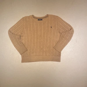 Polo Ralph Lauren Cableknit - Kabelstickad beige tröja från Polo Ralph Lauren med rund halsringning och klassisk broderad logga på bröstet. Tröjan har långa ärmar och ribbade muddar, perfekt för lager-på-lager under höst och vinter. Helt och hållet fläck och hålfri.