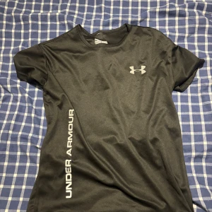 Svart Under Armour t-shirt i polyester - Svart t-shirt från Under Armour med logga på bröstet och text längs sidan. Tillverkad i lätt och snabbtorkande polyester, perfekt för träning. Klassisk passform och rund halsringning. Snygg och sportig stil för dig som gillar att röra på dig.