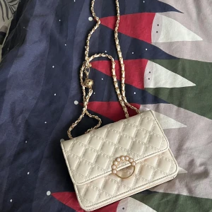 Quiltad beige axelväska med pärldetalj - Snygg beige axelväska med quiltat mönster och guldfärgad kedja. Väskan har ett dekorativt spänne med pärlor framtill och stilren, rektangulär form. Perfekt för dig som vill ha en trendig och elegant accessoar till din outfit.