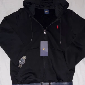Svart Ralph Lauren full zip hoodie  - Svart hoodie från Polo Ralph Lauren med dragkedja och huva. Klassisk röd broderad logga på bröstet och justerbara snören i huvan. Tillverkad i mjuk bomull, perfekt för chill dagar. Snygg och enkel att matcha med allt.