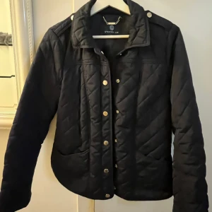 Svart quiltad jacka från Stockh LM - Snygg svart quiltad jacka från Stockh LM med guldiga knappar och axelremmar. Jackan har hög krage, två fickor framtill och markerade sömmar i rutmönster. Perfekt för dig som gillar stilren och klassisk look.