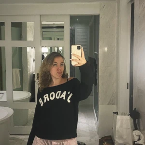 Svart stickad tröja med J'ADORE-tryck - Svart stickad tröja med vit text 'J'ADORE' framtill. Tröjan har en loose passform och bärs offshoulder för en avslappnad vibe. Långärmad och mjuk, perfekt för kyligare dagar. Enkel att styla med både jeans och leggings.