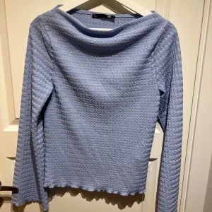 Ljusblå långärmad topp från 157 - Ljusblå långärmad topp från 157 med vågig struktur och lätt rullad kant nertill. Toppen har en lösare halsringning och är tillverkad i ett mjukt, stretchigt material som känns skönt mot huden. Perfekt för dig som gillar en chill och stilren look.