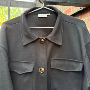 Höstjackan, svart, med två bröstfickor. -  klassisk krage, två stora bröstfickor med lock och snygga knappar framtill. Jackan har en rak och något loose passform, perfekt att slänga över en hoodie eller t-shirt. Materialet känns mjukt och har en clean look.