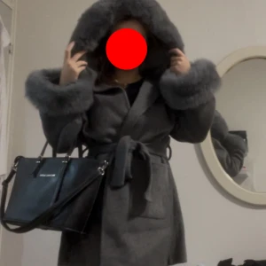 Grå kappa med päls och huva - Säljer en helt oanvänd snygg grå kappa med stor huva och breda fuskpälsdetaljer på ärmslut och huva. Kappan har bälte i midjan och två fickor framtill. Den är lite oversized så skulle säga att den passar folk med strl M eller L men det beror helt på preferenser. 