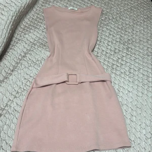 Ljusrosa klänning med bälte från Zara - Supersöt ljusrosa klänning från Zara med markerad midja och matchande bälte. Ärmlös och i kort modell, perfekt för varmare dagar. Enkel och stilren design som ger en feminin siluett.