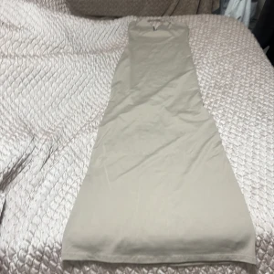 Beige långklänning med rosett från berska  - Supersnygg beige långklänning med smala halterneckband och liten svart rosett framtill. Klänningen har v-ringad urringning med svarta detaljer längs kanten och är figurnära upptill. Perfekt för dig som gillar stilrena och enkla plagg med en söt touch.