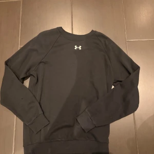 Svart sweatshirt från Under Armour - Svart långärmad sweatshirt från Under Armour med klassisk rund hals och logga på bröstet. Tröjan är i mjukt material och har ribbade muddar vid ärmslut och nederkant. Perfekt för chill eller träning och vardagen.