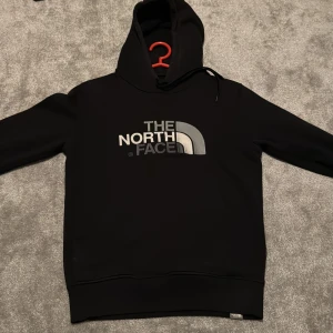 Svart hoodie från The North Face - Svart hoodie från The North Face med stor logga i vitt och grått på bröstet. Klassisk huva med snörning och ribbade muddar. Perfekt för dig som gillar streetwear och vill ha en stilren look. Materialet känns mjukt och bekvämt.