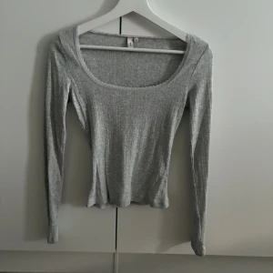 Grå ribbad långärmad topp - Säljer en grå ribbad långärmad topp från Nelly med rundad, djup urringning och diskret spetskant. Toppen är figurnära och har ett mjukt, stretchigt material som sitter snyggt på kroppen. Perfekt att styla med jeans eller kjol. Aldrig använd🩶