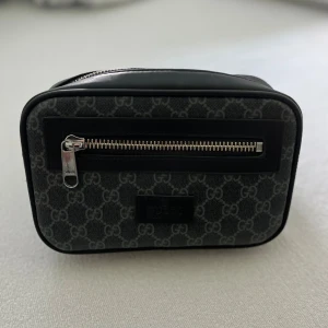 Svart waistbag med GG design - Snygg svart axelväska från G med klassiskt GG-mönster i grått. Väskan har en rektangulär form, dragkedja framtill och detaljer i skinn. Perfekt för dig som vill ha en stilren och lyxig accessoar.