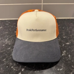 Peak Performance truckerkeps orange - Snygg truckerkeps från Peak Performance med vit front, mörkblå skärm och orange mesh på sidorna och bak. Klassisk böjd skärm och justerbar passform. Perfekt för dig som vill sticka ut med en sportig och cool look.