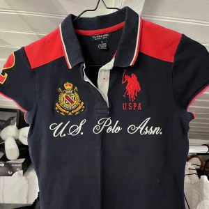 Mörkblå pikétröja från U.S. Polo Assn. - Snygg mörkblå pikétröja från U.S. Polo Assn. med röda axelpartier, broderad logga och emblem på bröstet samt vit text. Klassisk krage med knappar och korta ärmar med röda detaljer 🩷