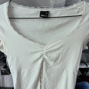 Vit topp med spets från Gina Tricot - Supersöt vit topp från Gina Tricot i storlek XS. Toppen har korta ärmar, v-ringning med spetskant och en snygg rynkdetalj framtill. Tillverkad i mjuk bomull som känns skön mot huden. Perfekt för dig som gillar en feminin och enkel stil. Aldrig andvänt! 🩷