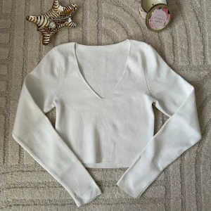 Vit ribbad croppad långärmad topp - Säljer en stilren vit croppad topp med lång ärm och djup v-ringning. Toppen är ribbad och har en tight passform som framhäver siluetten. Perfekt att matcha med jeans eller kjol för en clean look.