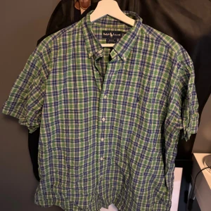 Rutig kortärmad skjorta Ralph Lauren XL - Kortärmad skjorta från Ralph Lauren i blått, grönt och vitt rutigt mönster. Klassisk button-down krage och knäppning framtill. Tillverkad i 100% bomull, perfekt för en chill och stilren look.