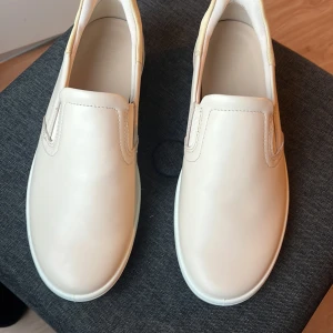 Rosa/krämvita slip-on sneakers från ECCO ny pris drygt 1200 - Stilrena vita slip-on sneakers från ECCO i slät skinn med elastiska paneler på sidorna för enkel påtagning. Skorna har rund tå, platt sula och diskret ECCO-logga under sulan. Perfekta för en clean och minimalistisk look. Skick 9/10 använd ca 3gånger 