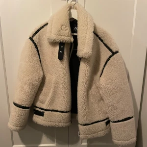 Vit teddyjacka från Zara - Säljer en vit teddyjacka från Zara med svarta detaljer och stora slag. Jackan har dragkedja, fickor och svarta band längs ärmar och midja. Väldigt varm och mysig, älskar till vintern!!