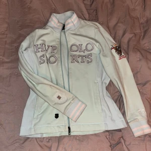 Ljusblå sportig jacka från HV Polo - Sportig ljusblå jacka från HV Polo med broderade loggor och emblem på bröstet och ärmen. Jackan har dragkedja framtill, ribbade muddar med ljusa ränder och hög krage. Perfekt för dig som gillar en stilren och preppy look. Strl 164 funkar som xs
