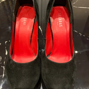 Svarta mockapumps med platå och röd sula - Snygga svarta pumps i mocka från Cesarre med hög klack och platå framtill. Skorna har en rund tå och insida samt sula i rött, vilket ger en riktigt cool kontrast. Perfekta för dig som vill sticka ut med en klassisk men edgy look.