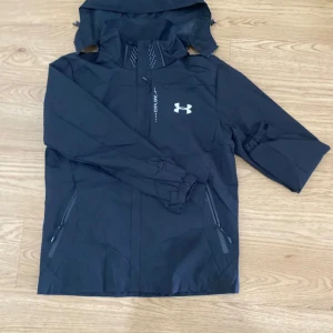 Svart vindjacka från Under Armour - Svart vindjacka från Under Armour med huva, dragkedjor på fickorna och halv dragkedja framtill. Jackan har elastiska muddar vid ärmsluten och är tillverkad i lätt syntetmaterial. Perfekt för dig som gillar sportig stil och vill ha något funktionellt.