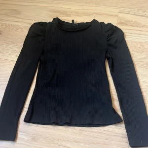 Svart ribbad långärmad topp Vero Moda - Svart långärmad topp från Vero Moda i ribbat material. Toppen har puffade axlar och en rund halsringning som ger en trendig siluett. Perfekt att styla med jeans eller kjol för en cool look.