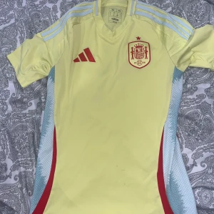 Spaniens fotbollströja Adidas XS gul - Gul Spanien fotbollströja från Adidas i slim fit, med röda detaljer och klubbmärke på bröstet. Tröjan har ljusblå ränder på axlarna och röda inslag på sidorna. Tillverkad i polyester, kortärmad och perfekt för match eller träning.