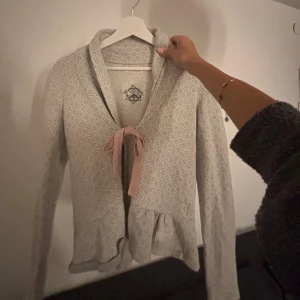 Odd Molly kofta med mönster och rosett - Supersöt ljusbeige kofta från Odd Molly med diskret mönster, volangkant nertill och rosa knytband framtill. Långärmad och perfekt för lager-på-lager. Mysig och feminin stil som lyfter vilken outfit som helst.