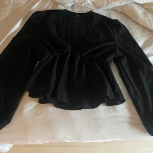 Svart blus med spetsdetaljer och v-ringning - Supersnygg svart blus med v-ringning och fina spetsdetaljer vid halsen. Blusen har långa puffärmar och markerad midja som ger en feminin siluett. Perfekt att styla med jeans eller kjol för en trendig look.