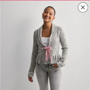 Grå mönstrad kofta med rosa knytband - Superfin grå kofta med vitt geometriskt mönster och volangkant nertill. Koftan har lång ärm, sjalkrage och ett gulligt rosa knytband i midjan. Koftan är i väldigt bra skick då den har blivit väldigt sällan använd