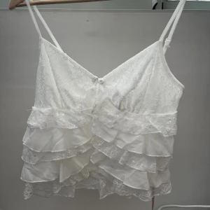 Vit volangig spetslinne med smala band - Supersöt vit linnetopp med flera lager volanger i spets och tunt tyg. Toppen har smala, justerbara axelband och v-ringning framtill. Perfekt för dig som gillar romantisk och feminin stil. Spetsdetaljerna ger en extra touch av elegans.