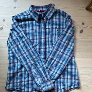 Rutig skjorta från Fjällräven M - Riktigt snygg rutig skjorta från Fjällräven i blått, rött och vitt. Skjortan har klassisk krage, bröstficka med broderad logga och knappar framtill. Perfekt för dig som gillar outdoor-vibbar och avslappnad stil.