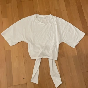 Vit croppad topp med knytband - Säljer en vit croppad topp med korta ärmar och långa knytband som kan knytas i ryggen. T-shirten har en lös passform och är tillverkad i mjuk bomull. Aldrig använt. Vid frågor och bild skriv gärna 🩷