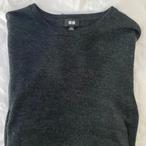 Mörkgrå stickad tröja från Uniqlo - En stilren mörkgrå stickad tröja från Uniqlo i klassisk modell. Tröjan har rund halsringning och långa ärmar, perfekt för lager-på-lager under kyliga dagar. Tillverkad i mjukt material som känns skönt mot huden.